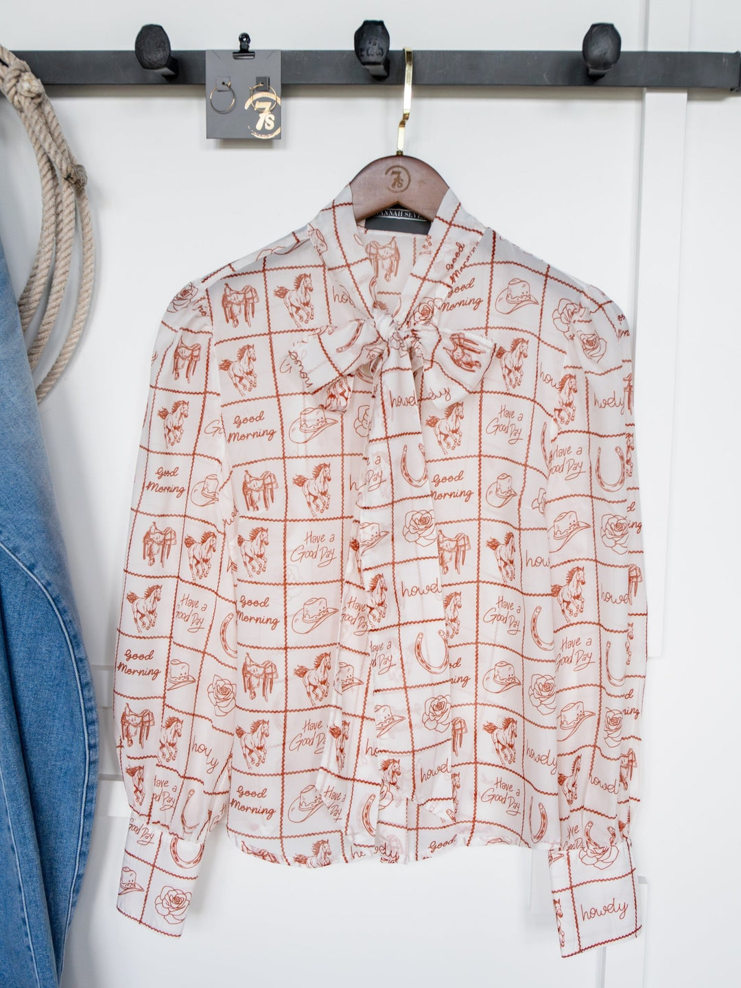The Good Day White Western Graphic Button Up Top by Savannah Sevens western life{&}style, featuring red grid patterns with animal and flower sketches and "Good Morning" text, hangs on a wooden hanger beside blue denim clothing.