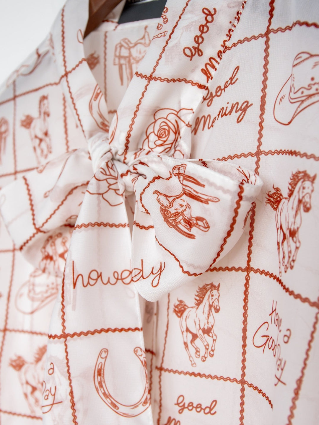 Close-up of the Good Day White Western Graphic Button Up Top by Savannah Sevens western life{&}style, a sheer blouse with a bold bow and Western prints—horses, cowboy hats, horseshoes, and phrases like "howdy" and "good morning.