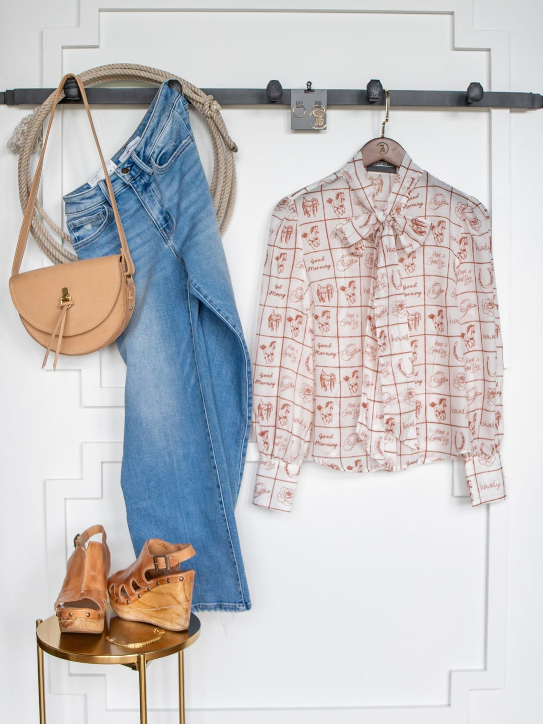 The Good Day White Western Graphic Button Up Top by Savannah Sevens western life{&}style is styled with jeans, a tan crossbody bag, and wedge sandals, all neatly displayed against a white wall with decorative molding.