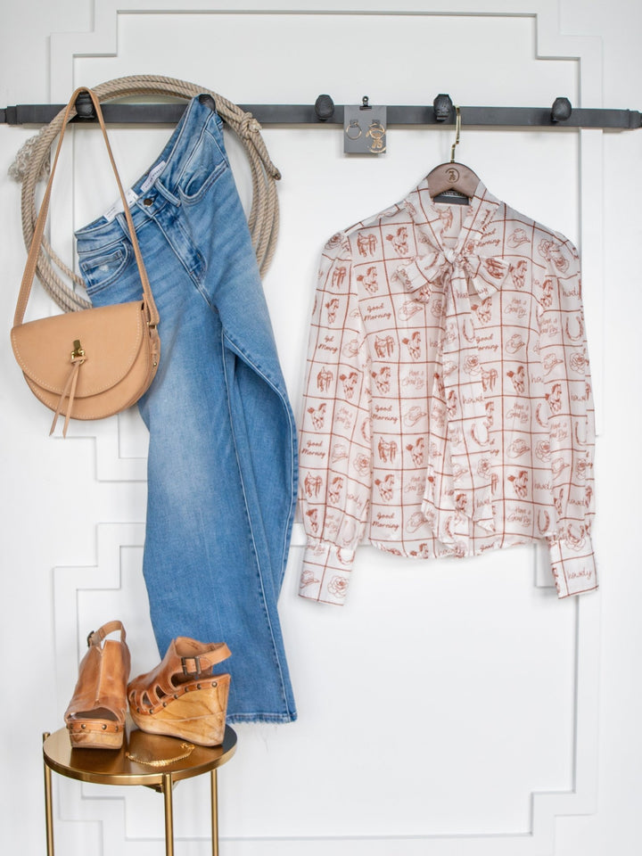 The Good Day White Western Graphic Button Up Top by Savannah Sevens western life{&}style is styled with jeans, a tan crossbody bag, and wedge sandals, all neatly displayed against a white wall with decorative molding.
