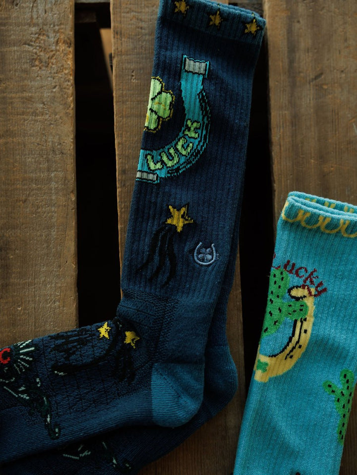 The Good Luck Crew Socks by Savannah Sevens western life{&}style, featuring dark blue with colorful horseshoe, stars, and "LUCKY" designs, are shown beside similar light blue performance socks on a wooden surface.