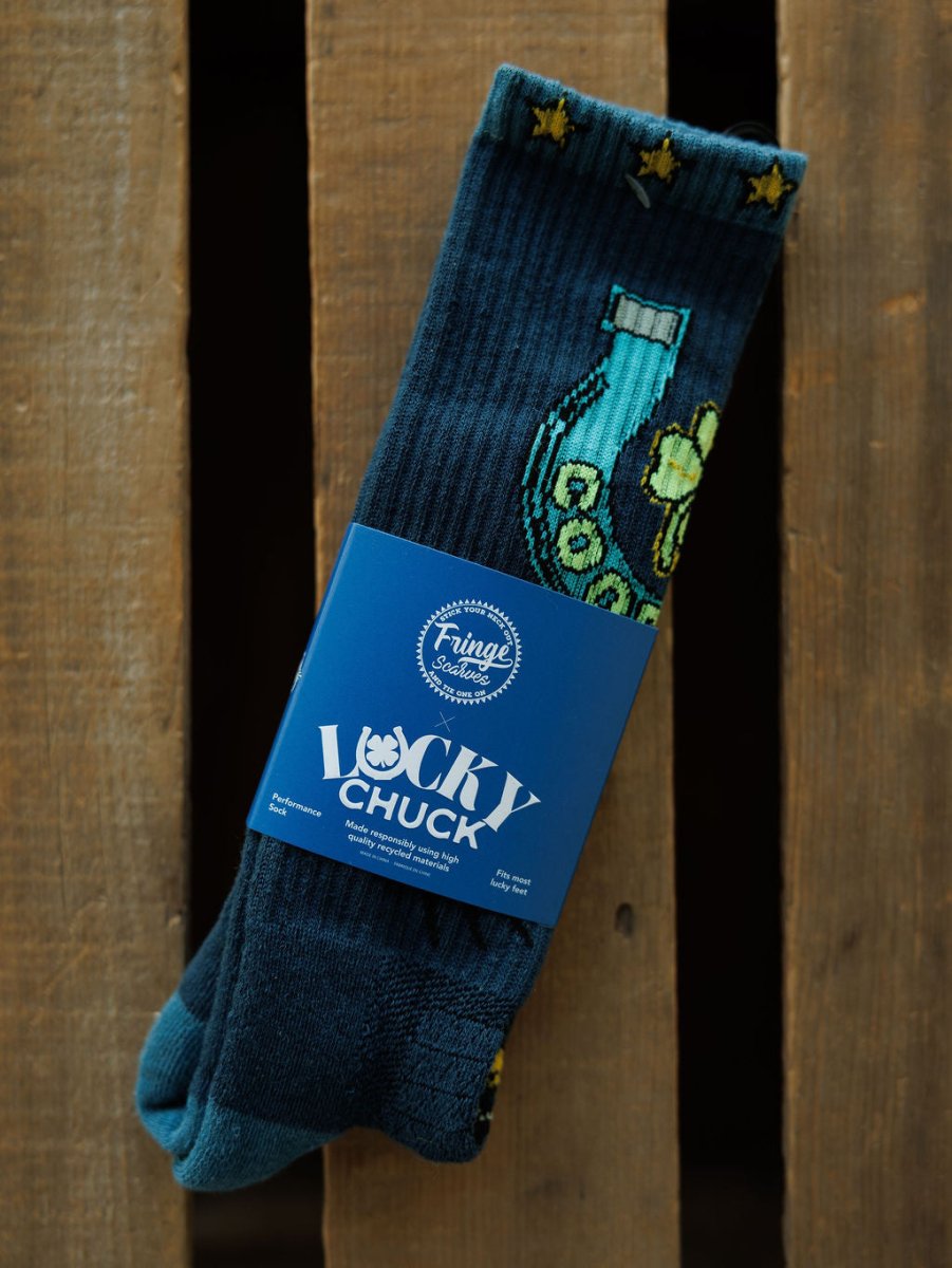 A pair of Good Luck Crew Socks by Savannah Sevens western life{&}style, featuring dark blue fabric with yellow stars, horseshoe and clover designs, wrapped in a blue label, is displayed on a wooden surface.