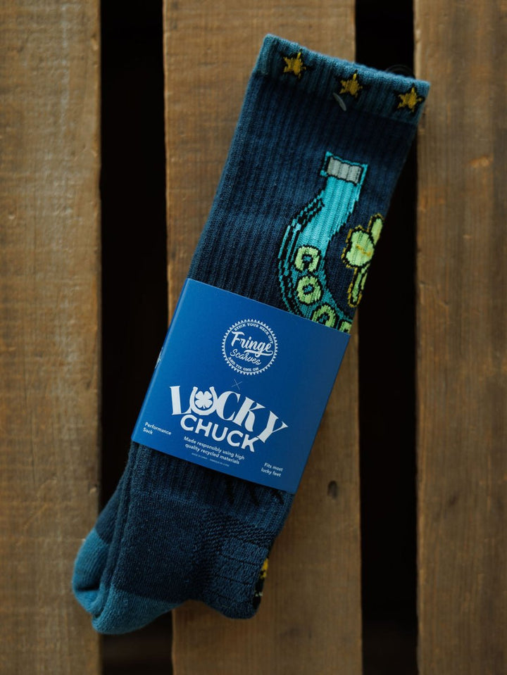 A pair of Good Luck Crew Socks by Savannah Sevens western life{&}style, featuring dark blue fabric with yellow stars, horseshoe and clover designs, wrapped in a blue label, is displayed on a wooden surface.
