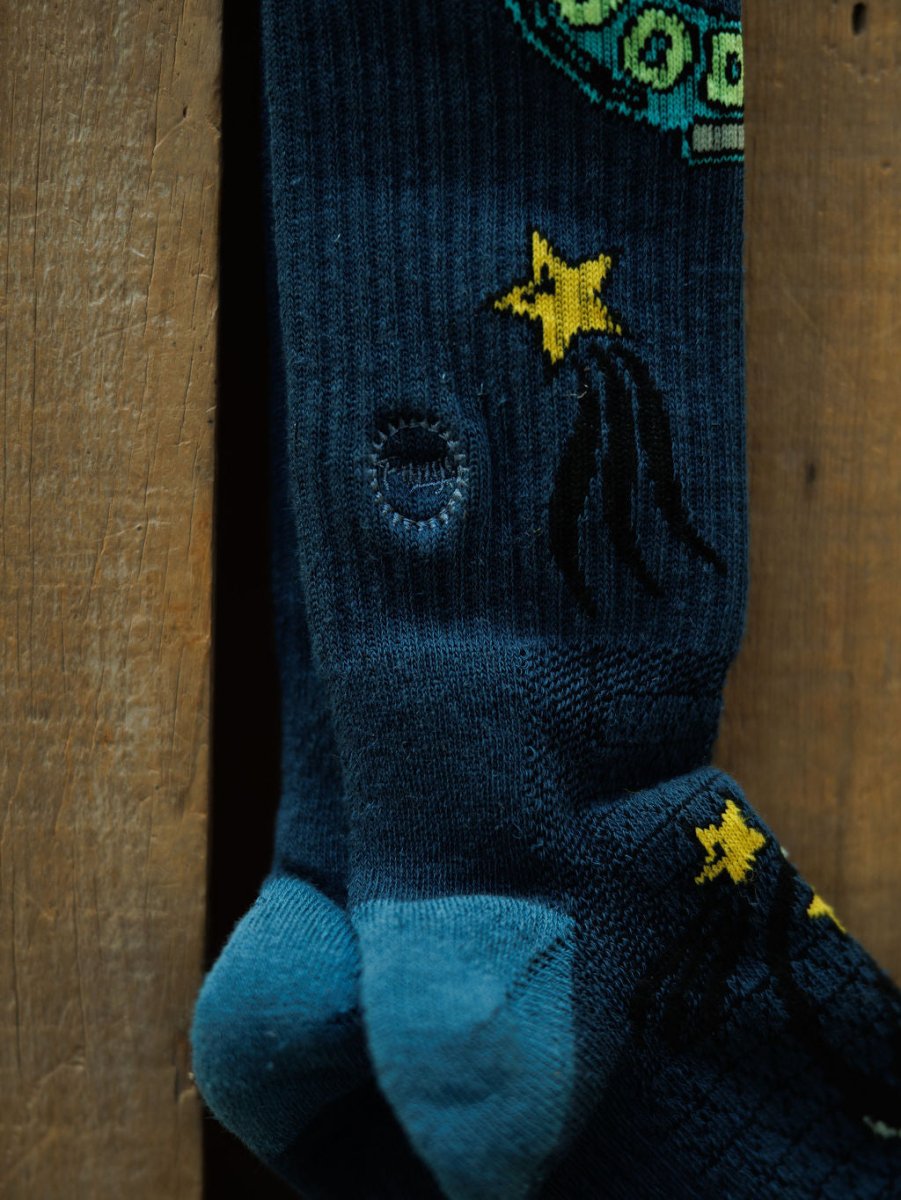 A close-up of Good Luck Crew Socks by Savannah Sevens western life{&}style in dark blue with a yellow star and black comet trail design, featuring a visible hole near the ankle, displayed on a wooden background.