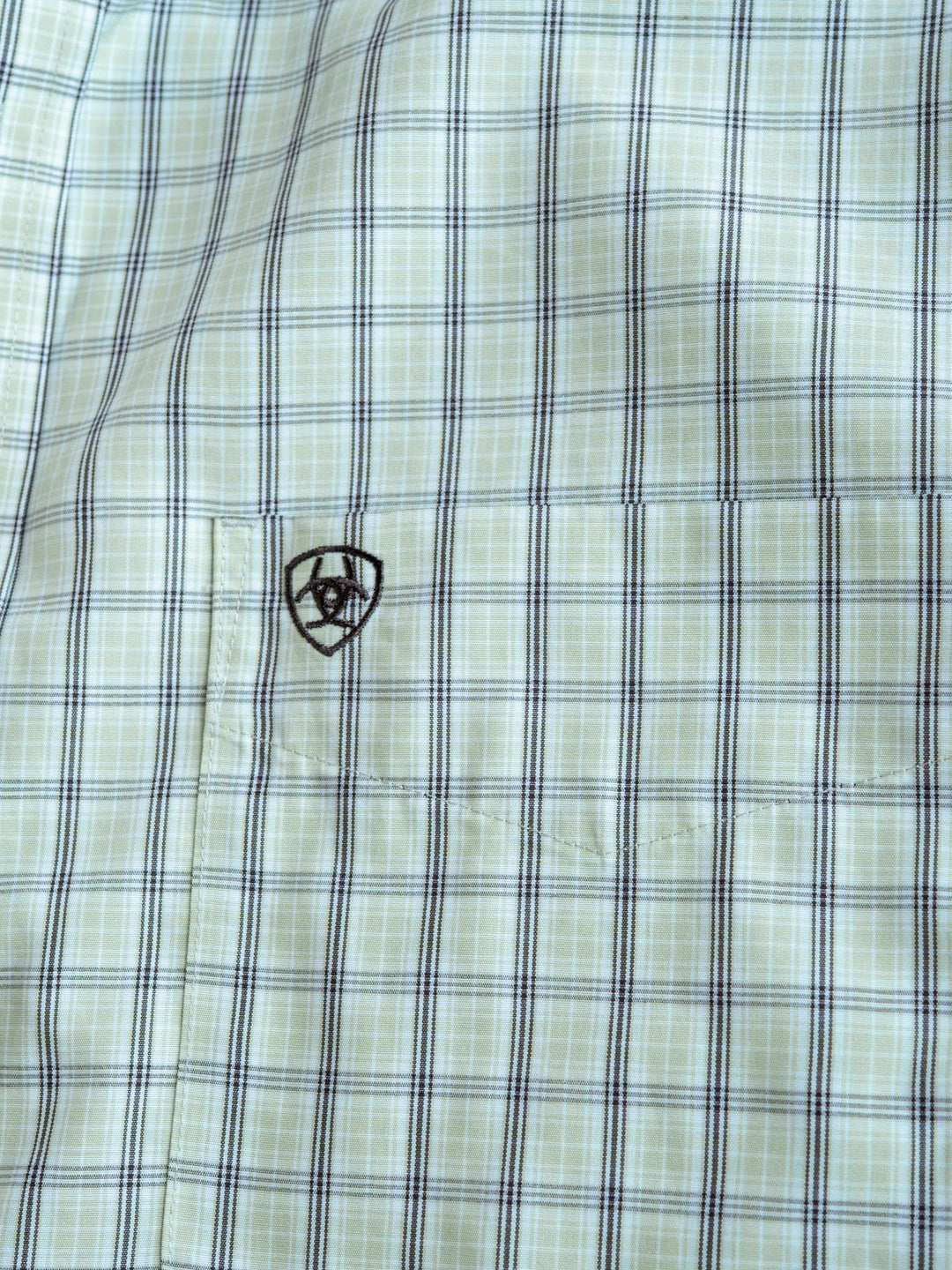 Close-up of the Graham Ariat Men's Plaid Button Up by Savannah Sevens western life{&}style, light green and blue, short sleeves, small front pocket with an embroidered black shield, horseshoe & star emblem. Wrinkle-free fabric for easy care.