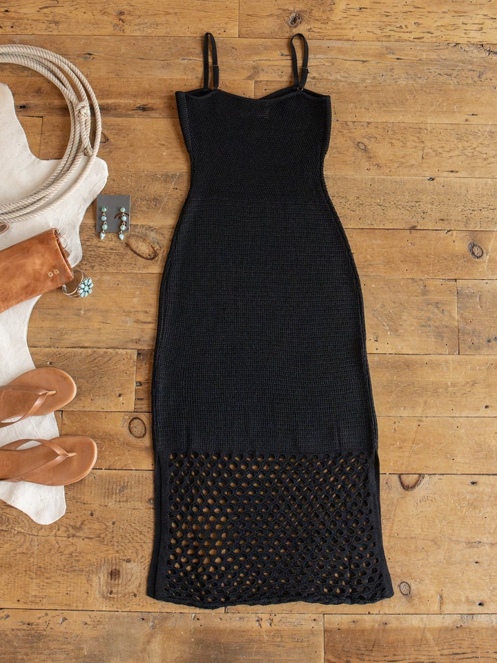 The Granada Black Crochet Dress by Savannah Sevens western life{&}style is displayed flat on a wooden floor, featuring a sleeveless design and mesh-patterned hem, and is styled with tan sandals, and turquoise earrings.