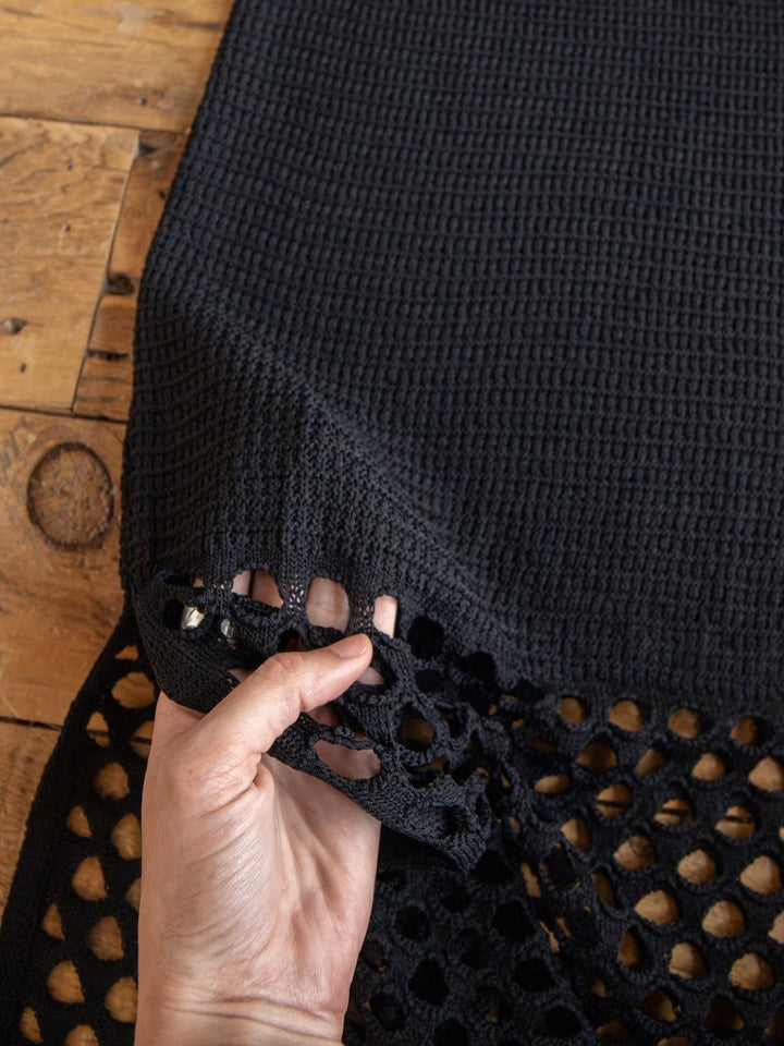 A hand lifts the edge of the Savannah Sevens western life{&}style Granada Black Crochet Dress, revealing its textured fabric with a grid-like open pattern along one side, displayed on a wooden surface.