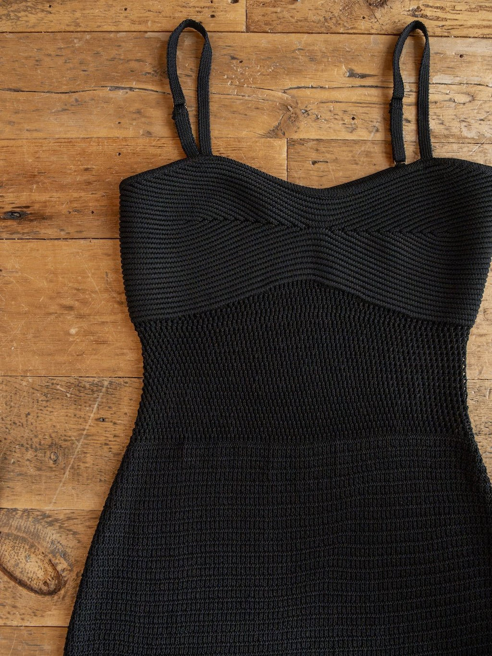 The Granada Black Crochet Dress by Savannah Sevens western life{&}style is shown lying flat on a wooden floor. Only the top half of the sleeveless, full-length dress is visible, highlighting its thin straps, structured bodice, and textured crochet fabric.