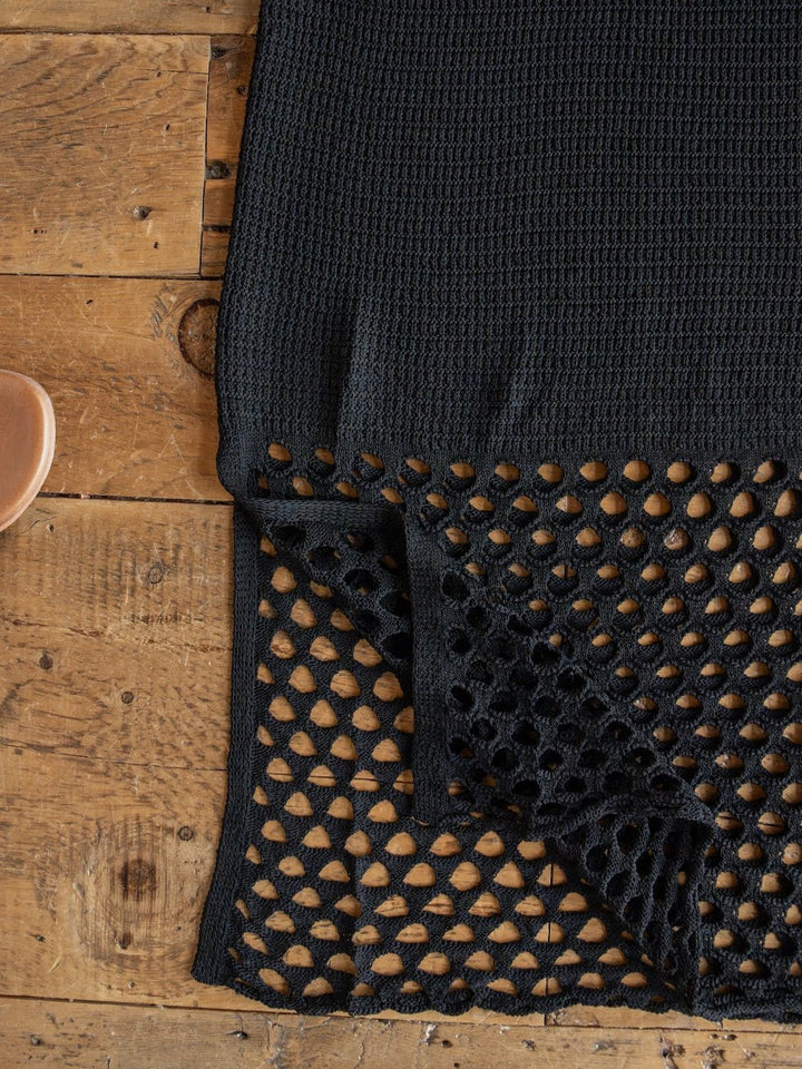 A black crochet fabric from the Granada Black Crochet Dress by Savannah Sevens western life{&}style is displayed on a rustic wooden surface, with one corner folded over to reveal its intricate mesh-edge detail—evoking the sheer accents of its v-neck design.