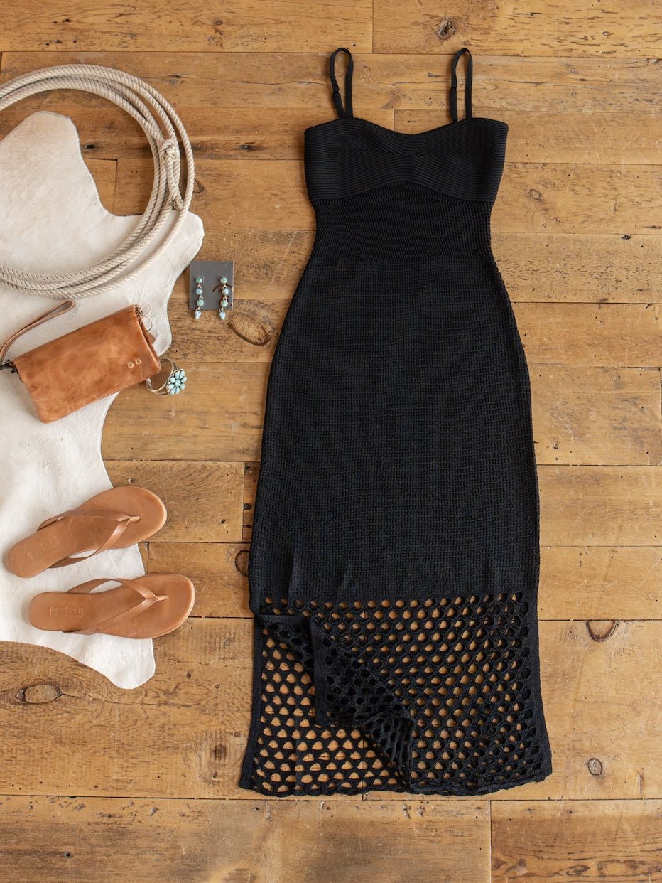 The Granada Black Crochet Dress from Savannah Sevens western life{&}style, a black sleeveless full-length piece featuring a mesh hem, is displayed on a wooden floor alongside tan sandals, a brown bag, turquoise earrings, and a white cowhide rug.