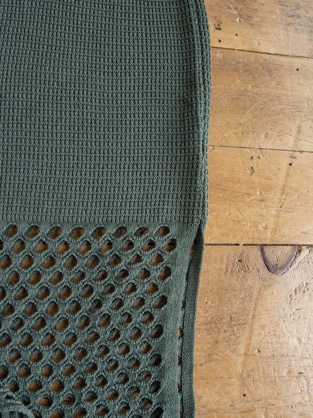 A close-up of the textured green fabric from the Savannah Sevens western life{&}style Granada Blue Sage Crochet Dress, showing a sleeveless full-length design with a waffle pattern on top and a larger open mesh pattern below, laid out on a wooden surface.