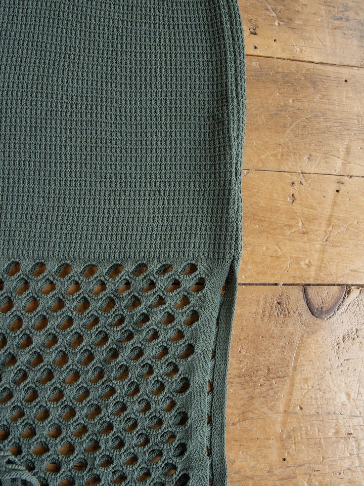 A close-up of the textured green fabric from the Savannah Sevens western life{&}style Granada Blue Sage Crochet Dress, showing a sleeveless full-length design with a waffle pattern on top and a larger open mesh pattern below, laid out on a wooden surface.