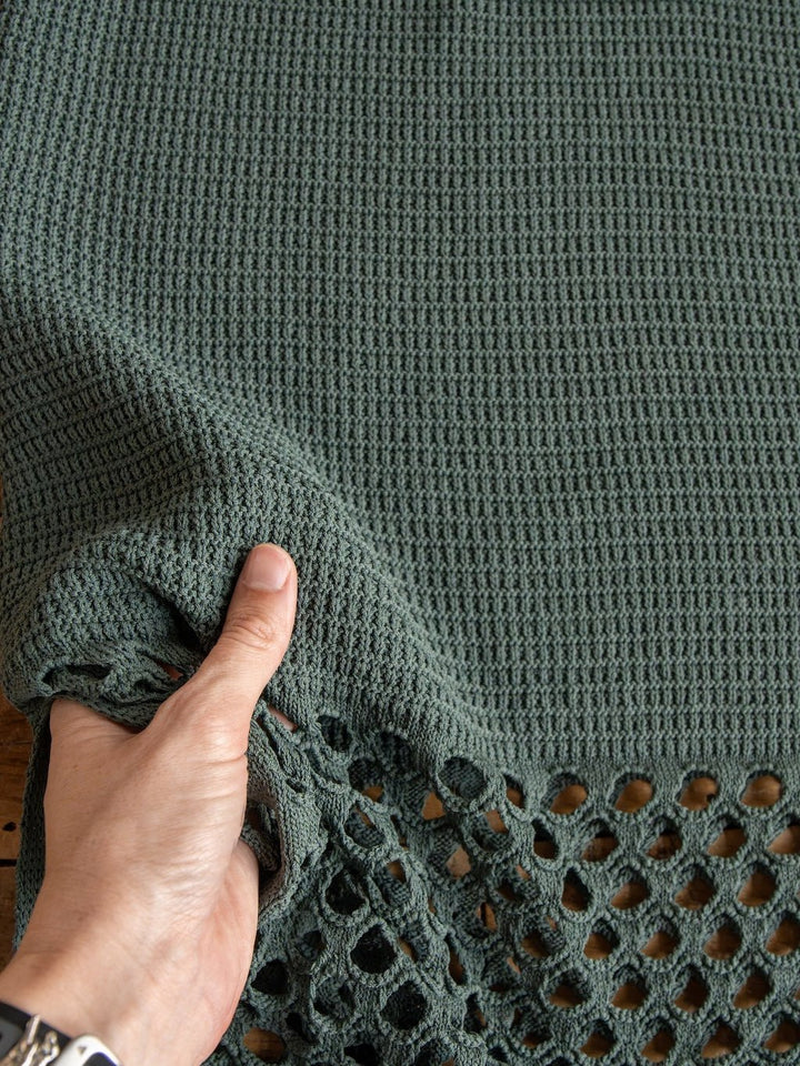 A hand holds the edge of a green, waffle-textured fabric with an open, airy crochet section at the bottom—ideal for crafting the Savannah Sevens western life{&}style Granada Blue Sage Crochet Dress, a sleeveless full-length design with petite adjustable straps.