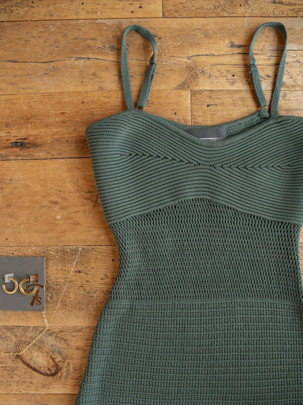 A Granada Blue Sage Crochet Dress by Savannah Sevens western life{&}style, featuring a textured green design, sleeveless full-length silhouette, and petite adjustable straps, is laid out on a wooden floor next to a pair of gold hoop earrings and a gold chain bracelet.