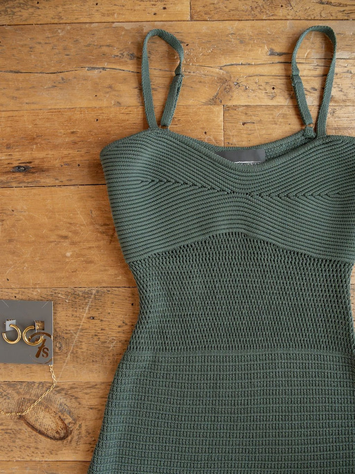 A Granada Blue Sage Crochet Dress by Savannah Sevens western life{&}style, featuring a textured green design, sleeveless full-length silhouette, and petite adjustable straps, is laid out on a wooden floor next to a pair of gold hoop earrings and a gold chain bracelet.