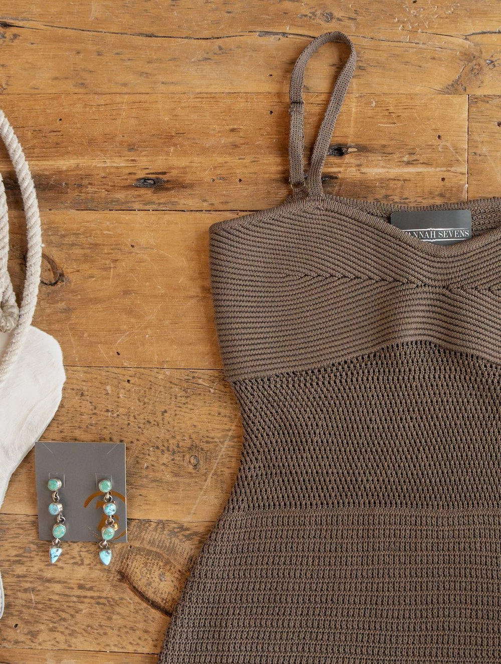 The Granada Chocolate Crochet Dress by Savannah Sevens western life{&}style, featuring petite adjustable straps, is displayed on a wooden surface. Beside it are a pair of dangling turquoise earrings and a cream-colored bag with a rope handle.