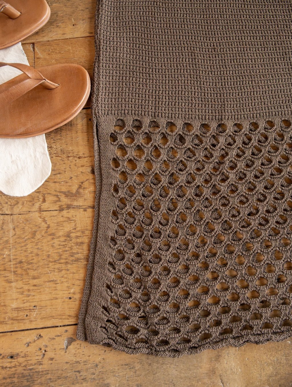The Granada Chocolate Crochet Dress from Savannah Sevens western life{&}style, featuring an open-weave crochet pattern at the hem, is displayed on a wooden floor alongside tan sandals —highlighting the garment’s intricate, handcrafted style.