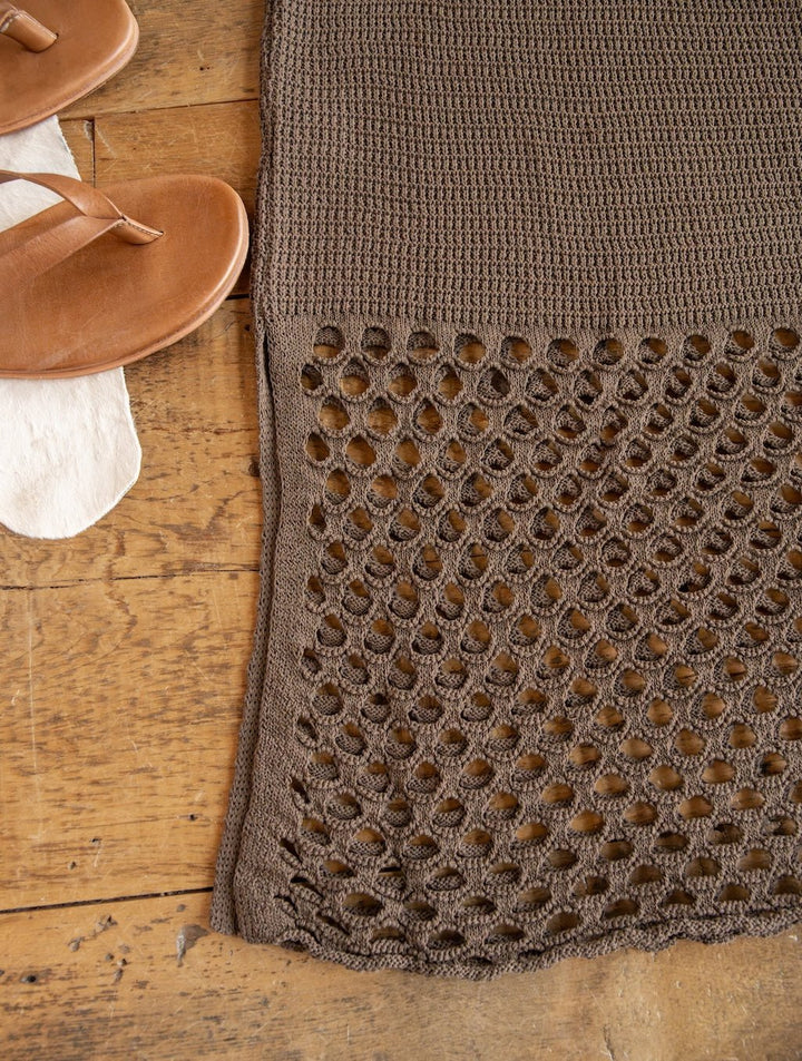 The Granada Chocolate Crochet Dress from Savannah Sevens western life{&}style, featuring an open-weave crochet pattern at the hem, is displayed on a wooden floor alongside tan sandals —highlighting the garment’s intricate, handcrafted style.