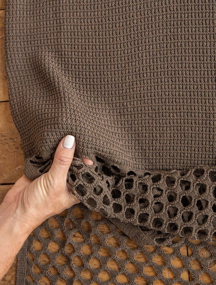 A hand  holds the Granada Chocolate Crochet Dress by Savannah Sevens western life{&}style, showcasing its textured brown crochet fabric and open, honeycomb-like details over a wooden surface.
