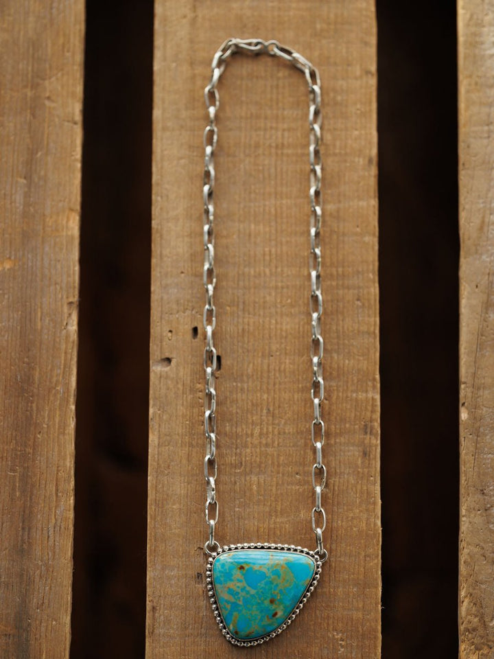 The Granada Necklace by Savannah Sevens western life{&}style features a large triangular Kingman turquoise pendant with brownish patterns on sterling silver, displayed on a rustic wooden surface.