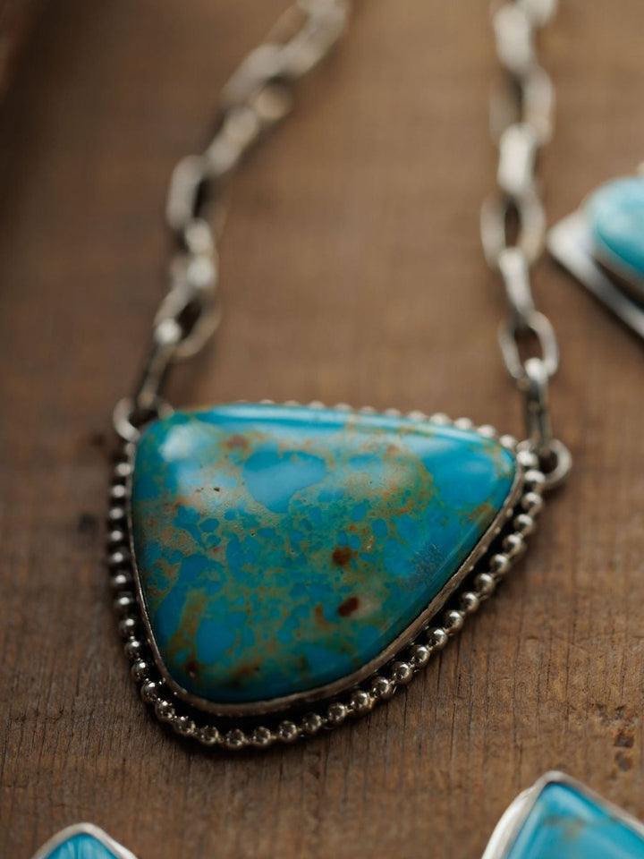 A close-up of the Granada Necklace by Savannah Sevens western life{&}style, featuring Kingman turquoise with brown and green patterns in a sterling silver beaded frame on a silver chain, resting on wood.