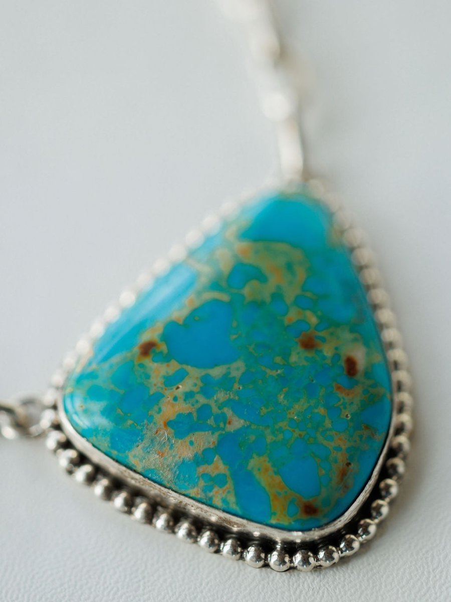 A close-up of the Granada Necklace by Savannah Sevens western life{&}style, featuring a triangular Kingman turquoise pendant with brown and green speckles in a sterling silver beaded frame, shown on a light background.