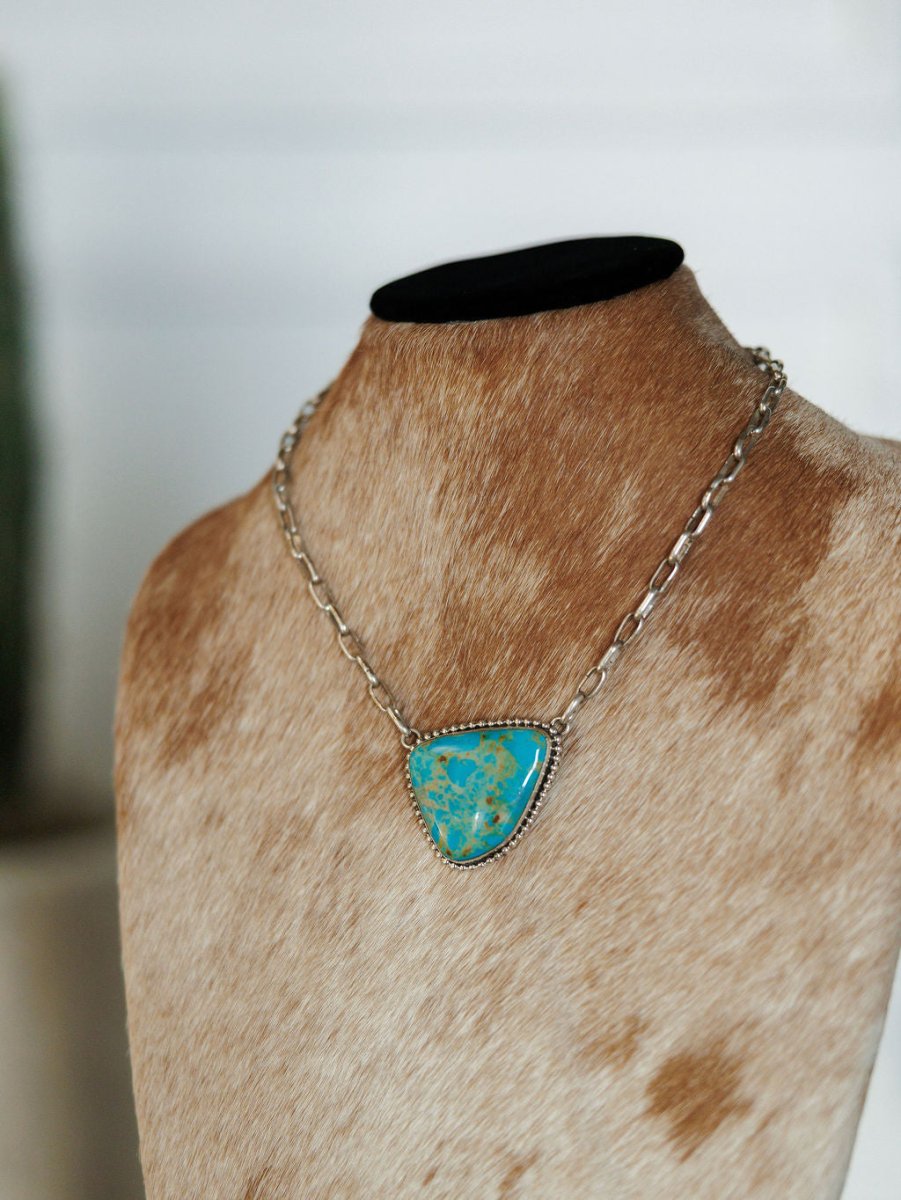The Granada Necklace by Savannah Sevens western life{&}style is a sterling silver chain with a triangular Kingman turquoise pendant, shown on a brown and white cowhide mannequin bust.