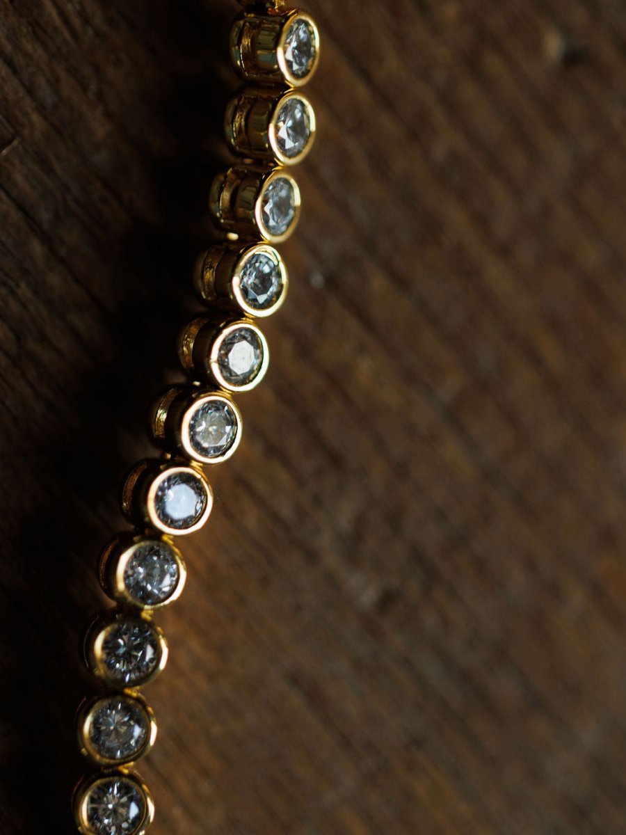 A close-up of the Granby Bracelet by Savannah Sevens western life{&}style, featuring round clear cubic zirconia stones set in 18K gold plating, displayed on a textured wooden surface with a blurred background.