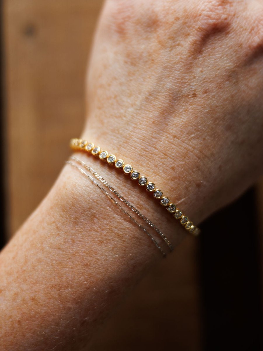A close-up of a wrist wearing the Granby Bracelet by Savannah Sevens western life{&}style—an 18K gold plated, hypoallergenic piece with round gemstones, paired with minimalist silver chain bracelets against a blurred wood background.