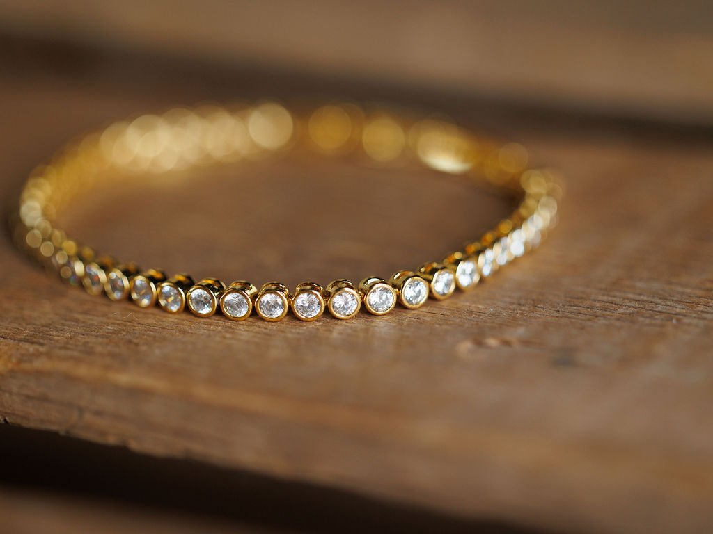 The Granby Bracelet by Savannah Sevens western life{&}style features round, clear cubic zirconia stones set in a row on a gold tennis bracelet, displayed on wood with a blurred background.