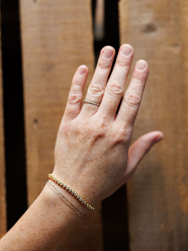 A hand wears the Granby Bracelet by Savannah Sevens western life{&}style—an 18K gold plated, hypoallergenic beaded bracelet—alongside a thin ring and two other bracelets, resting on a wooden surface with visible grain and board gaps.