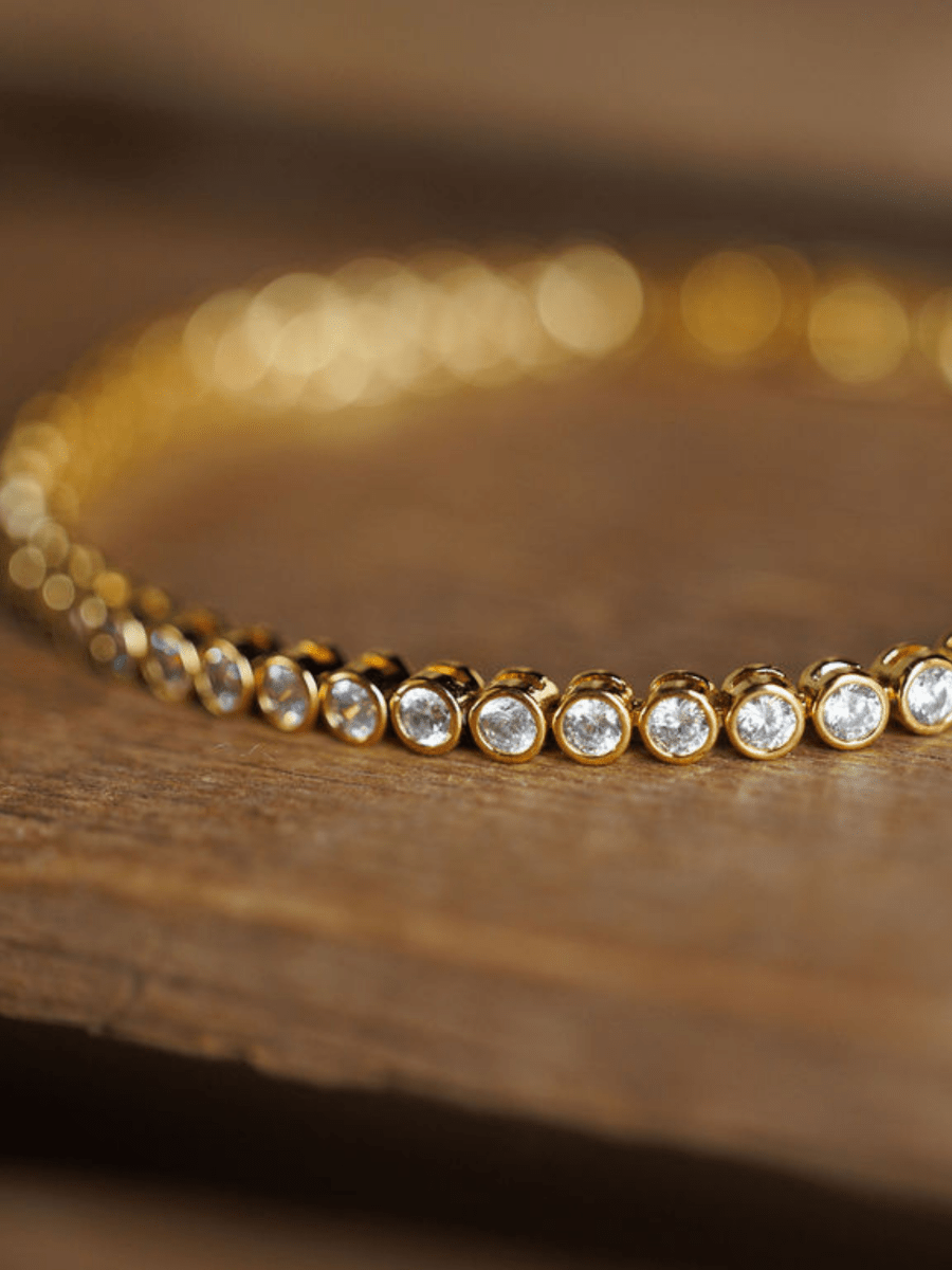 The Granby Bracelet by Savannah Sevens western life{&}style, an 18K gold plated piece with round bezel-set cubic zirconia stones, sparkles in focus on a wooden surface while the background is softly blurred.
