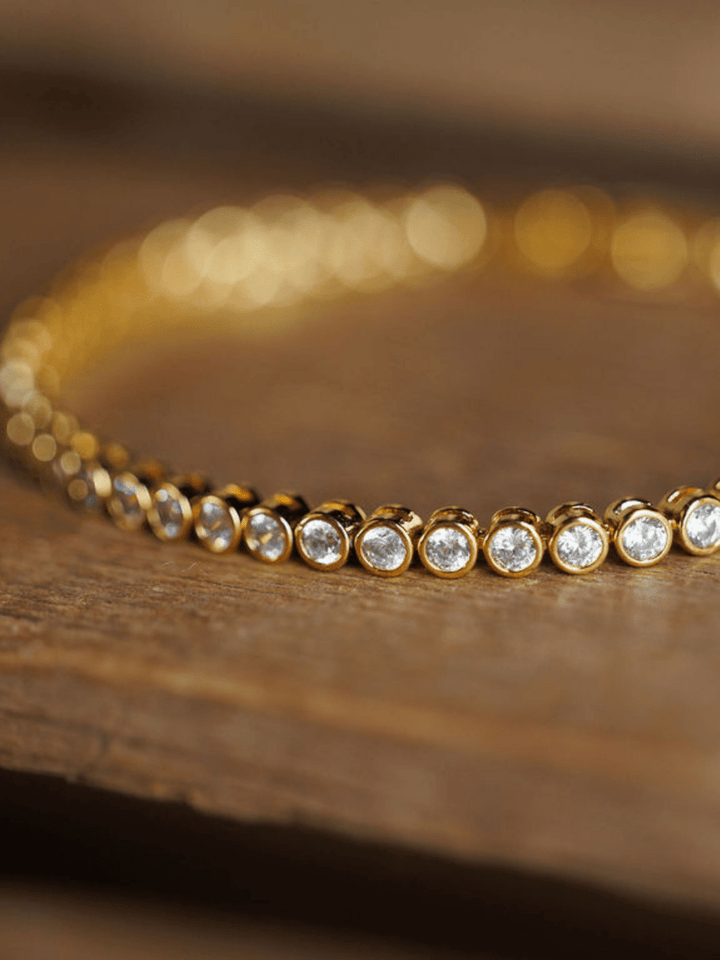 The Granby Bracelet by Savannah Sevens western life{&}style, an 18K gold plated piece with round bezel-set cubic zirconia stones, sparkles in focus on a wooden surface while the background is softly blurred.