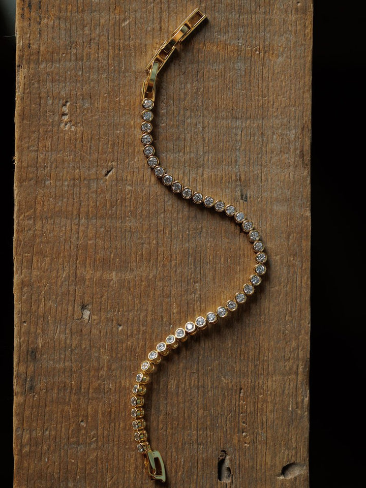 The Granby Bracelet by Savannah Sevens western life{&}style features round diamonds in a wavy line on an 18K gold-plated, hypoallergenic design. Shown on rustic wood, it offers elegant comfort for everyday wear.