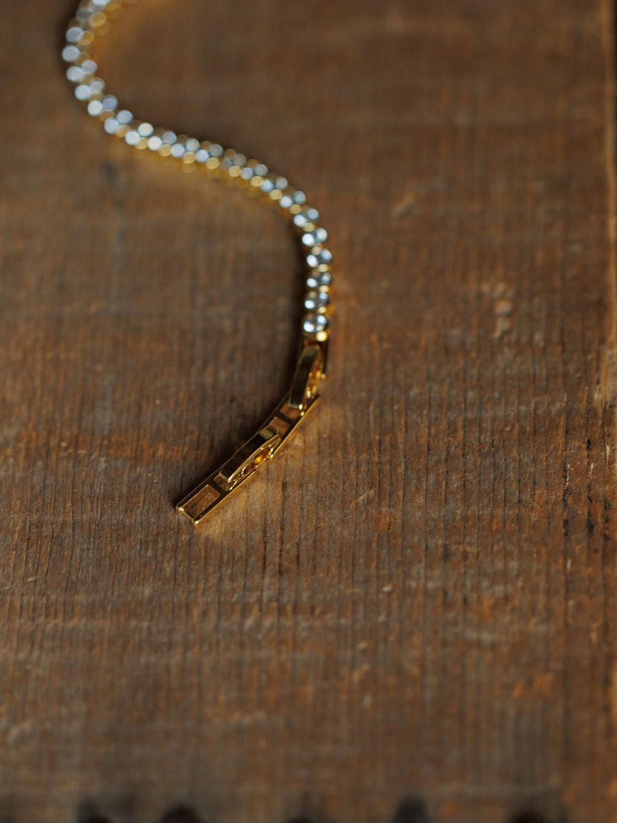 A close-up of the Granby Bracelet by Savannah Sevens western life{&}style, an 18K gold plated, hypoallergenic diamond tennis bracelet with a geometric clasp, shown on a textured wooden surface.