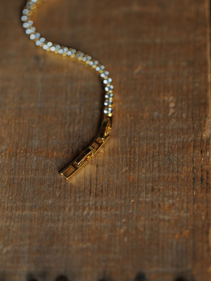 A close-up of the Granby Bracelet by Savannah Sevens western life{&}style, an 18K gold plated, hypoallergenic diamond tennis bracelet with a geometric clasp, shown on a textured wooden surface.
