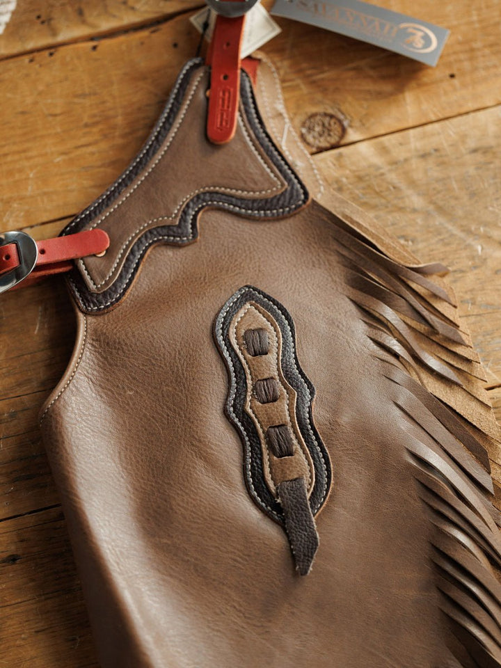 The Granger Chinks {Youth} by Savannah Sevens western life{&}style feature brown handmade leather with decorative stitching, fringe, red leather straps with buckles, and a branded tag, displayed on a wooden surface.