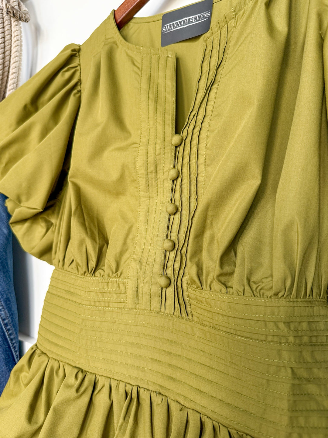 A close-up of the Grangeville Olive Puff Sleeve Top by Savannah Sevens western life{&}style, featuring pintuck details, fabric-covered buttons, and a pleated elastic waistline. This cotton blend blouse is displayed on a wooden hanger.