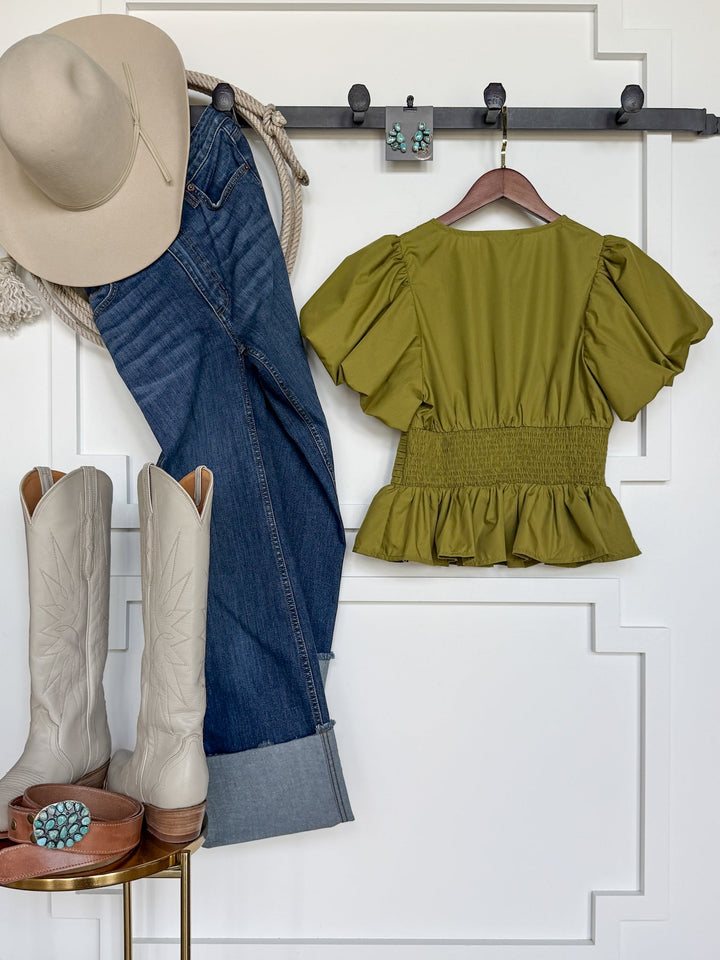 Western outfit laid out: beige cowboy hat, blue jeans, Grangeville Olive Puff Sleeve Top by Savannah Sevens western life{&}style (featuring an elastic waistline), white cowboy boots, brown belt with turquoise buckle, and turquoise earrings—all hanging or placed against a white decorative wall.
