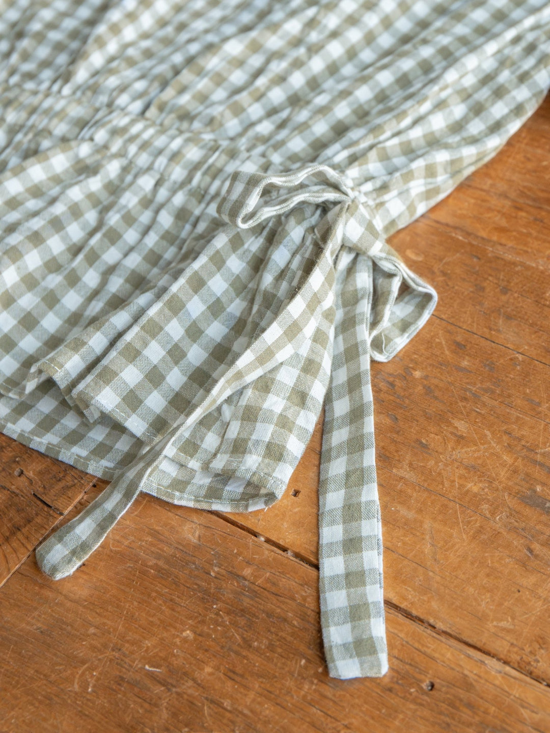 A close-up of The Grazen by Savannah Sevens western life{&}style—a sage green and white gingham top with a tied bow detail—resting on a wooden surface.