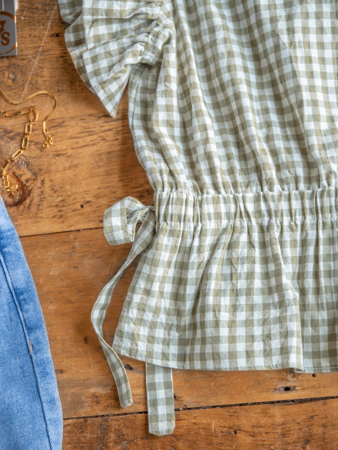 The Grazen by Savannah Sevens western life{&}style—a sage gingham short sleeve top with waist tie detail—is shown on a wooden surface beside light blue denim and a gold earring.