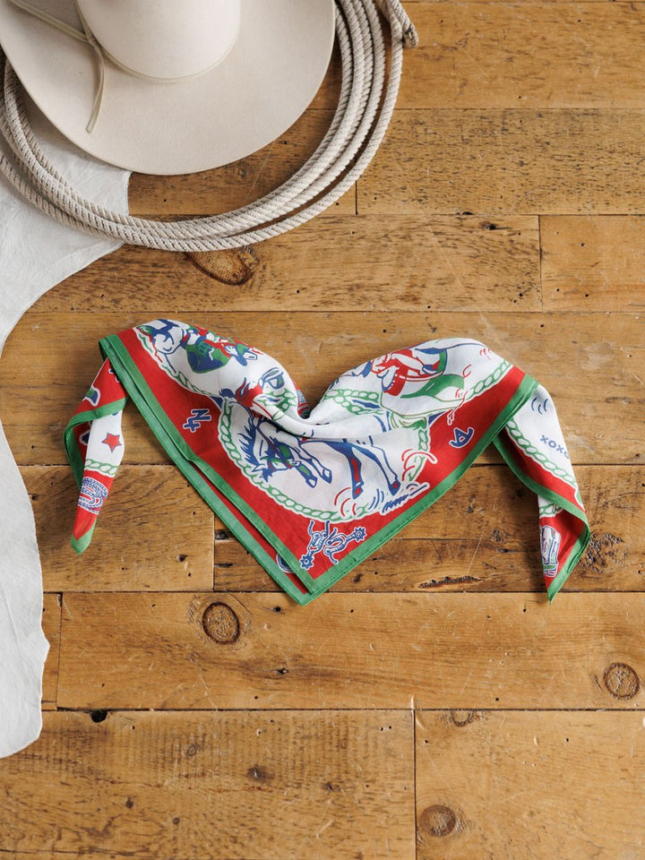 The Greetings Neck Scarf by Savannah Sevens western life{&}style, featuring green, red, and white designs, is folded on a wooden surface beside a white cowboy hat, rope, and a piece of white leather.