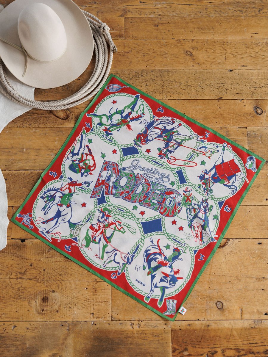 The Greetings Neck Scarf by Savannah Sevens western life{&}style, featuring vibrant cowboy and rodeo designs with a red and green border, is displayed on a wooden floor beside a white cowboy hat and coiled rope.