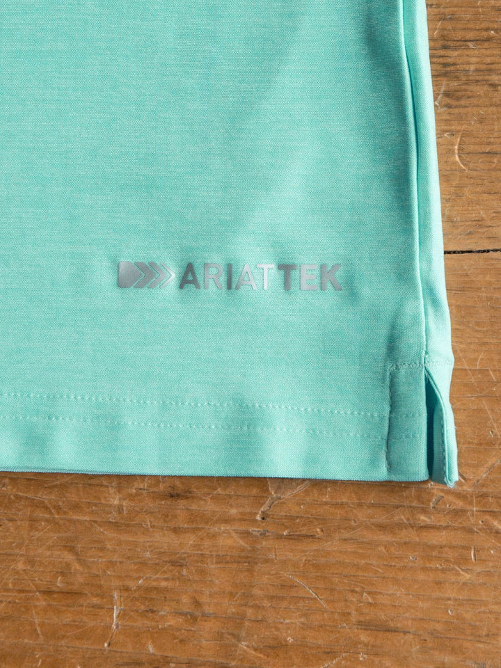 A close-up of Griffin Ariat Men's Turquoise Polo fabric by Savannah Sevens western life{&}style, featuring the silver ARIATTEK logo near the hem on a wooden surface. The polo offers moisture-wicking for comfort.