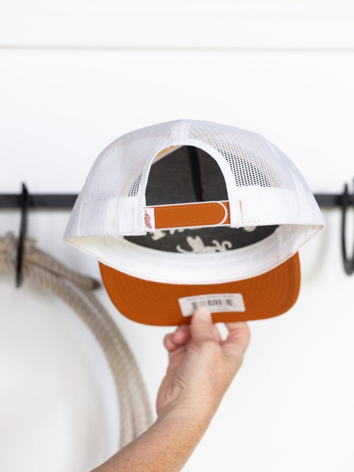 A hand holds up a Gruene Hall Cap by Savannah Sevens western life{&}style, featuring a white mesh back and burnt orange brim, showing the inside and underside in front of a white wall with black hooks.