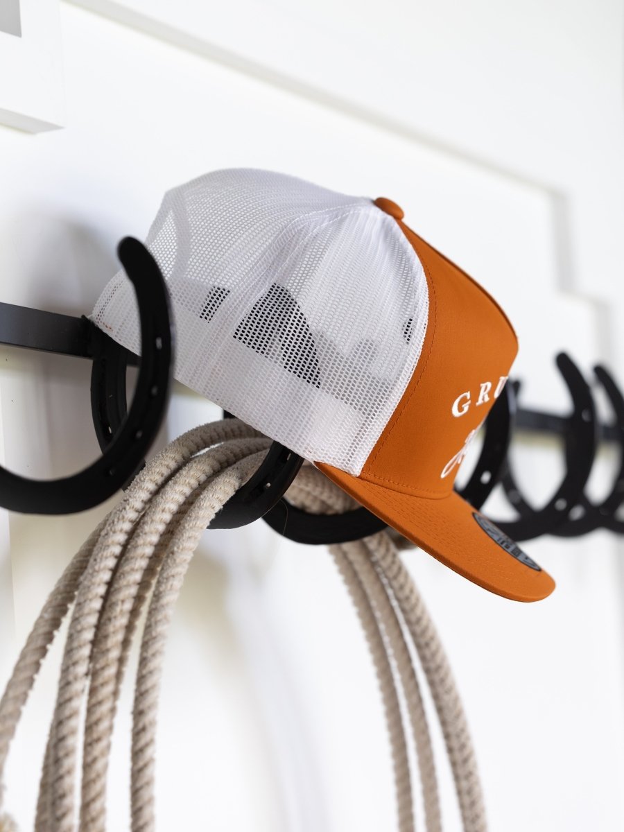 A Gruene Hall Cap by Savannah Sevens western life{&}style in orange and white sits on a black horseshoe-shaped wall rack next to a coiled rope, set against a white background.