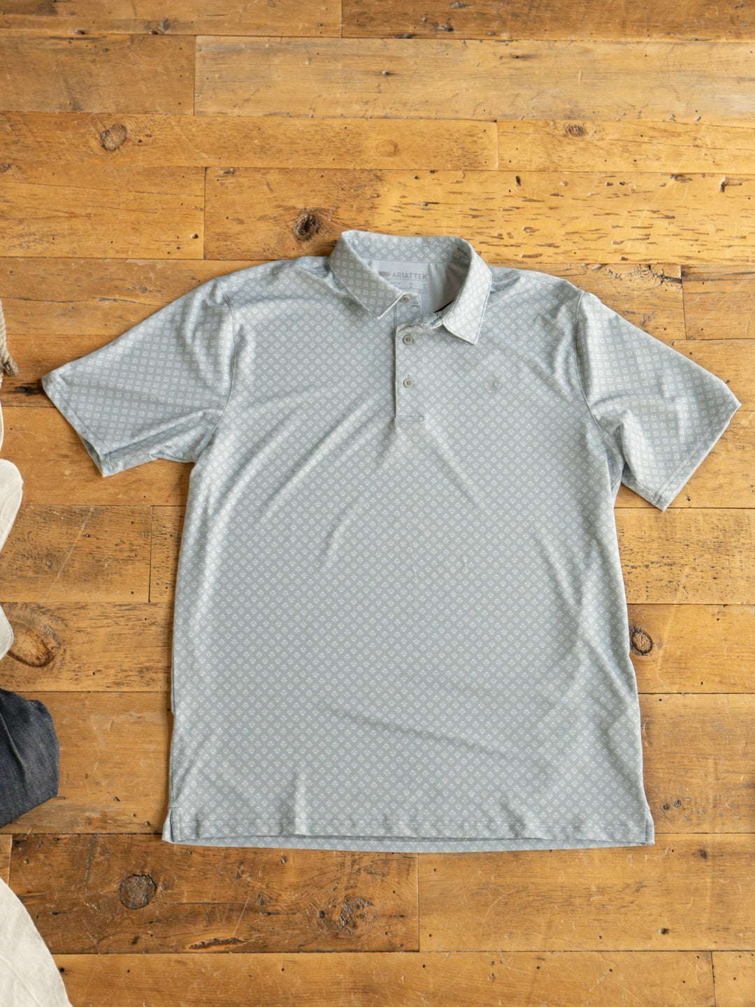 The Guymon Ariat Men's Southwest Polo by Savannah Sevens western life{&}style, in light gray with a subtle pattern, is laid flat on a wooden floor. A white garment and dark fabric appear near the edges. This breathable men’s polo offers all-day comfort and odor resistance.