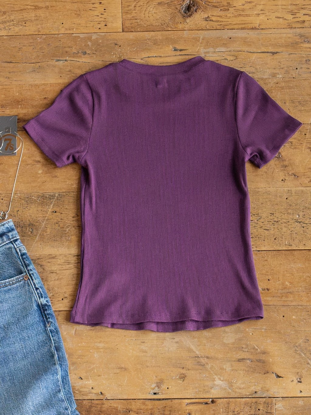 The Hadley Plum Ribbed Basic Tee by Savannah Sevens western life{&}style, a young contemporary fitted short-sleeve top, is laid flat on a wooden floor beside the top corner of a pair of blue jeans.