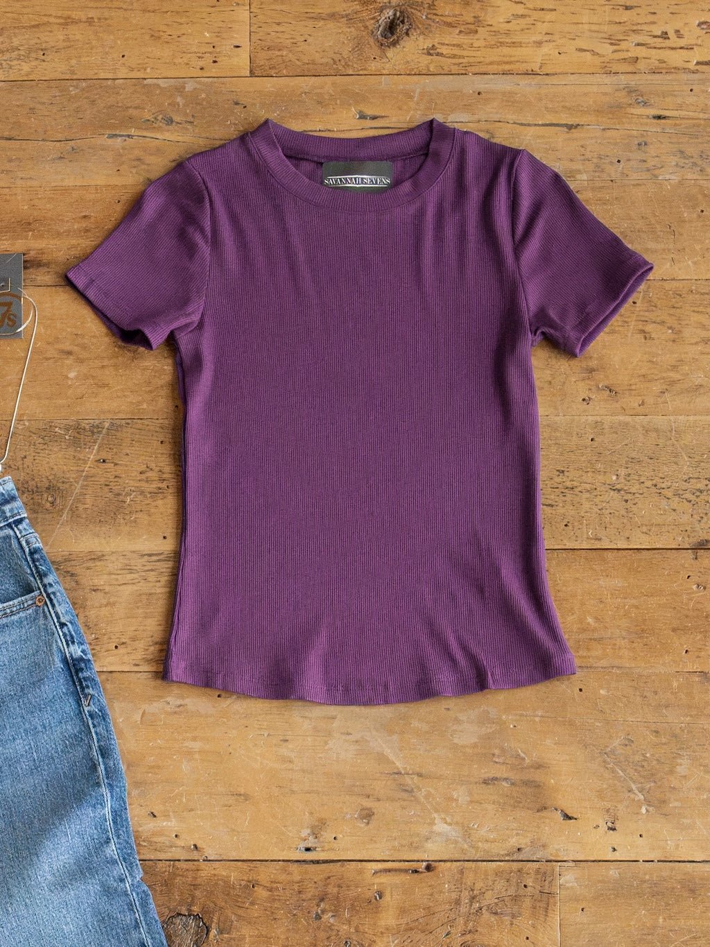 The Hadley Plum Ribbed Basic Tee by Savannah Sevens western life{&}style is displayed flat on a wooden floor beside the edge of a pair of blue jeans.