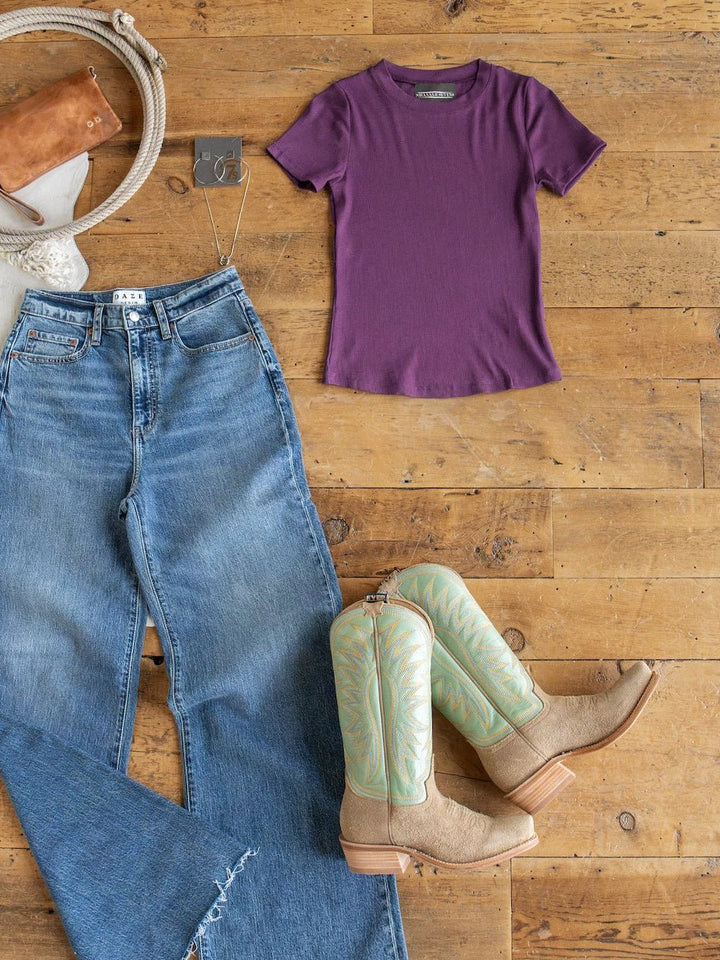 Flat lay of blue jeans, tan cowboy boots, a beige rope belt, a brown purse, silver earrings, and the Hadley Plum Ribbed Basic Tee by Savannah Sevens western life{&}style arranged on a wooden floor.